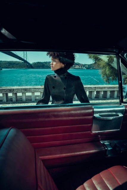 Stylish woman in a coat poses inside a classic car with a turquoise sea and bridge visible through the open window.