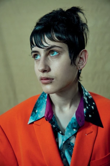 Close-up portrait of a young person with dark hair and lip piercing, wearing an orange blazer and galaxy-print shirt against neutral backdrop.