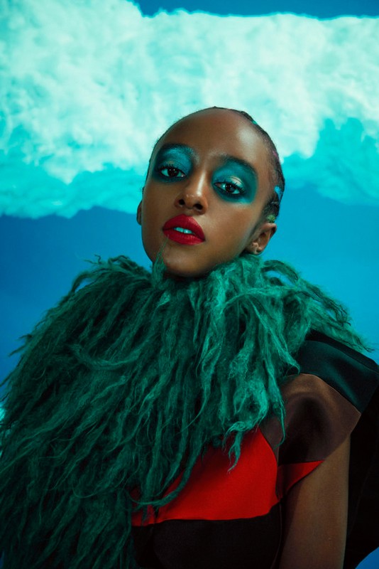 Portrait of an African-descent woman wearing bright blue eyeshadow and red lipstick, a green fringe shawl, with a cloudy blue backdrop.