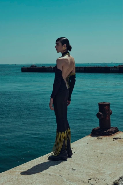 A woman in a black dress stands on a concrete pier, gazing out at a tranquil, aquamarine ocean under a clear blue sky.