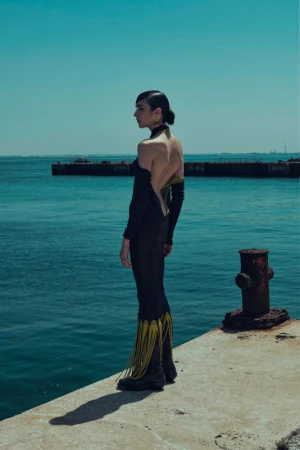 A woman in a black dress stands on a concrete pier, gazing out at a tranquil, aquamarine ocean under a clear blue sky.