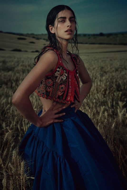 Woman poses in a field of wheat wearing a decorative top and blue skirt. Dark, moody lighting emphasizes her eyes and the natural setting.