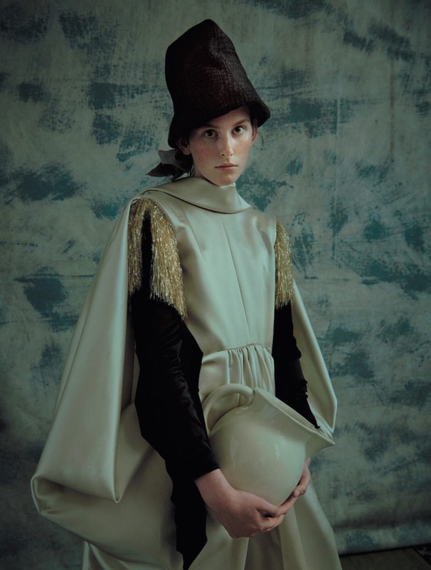 A person in a pale green gown and tall dark hat holds a white pitcher against a mottled blue background in a fashion portrait.