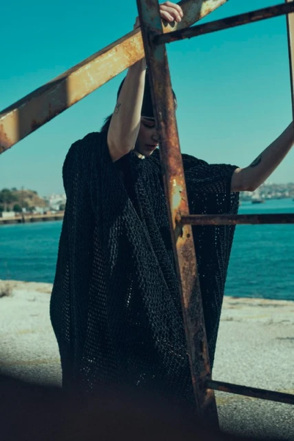 An adult female wearing a black netted poncho stands on a concrete structure, holding onto a rusty metal ladder with the sea in the background.