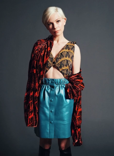 Fashionable woman in patterned top and blue leather skirt poses in a studio with a neutral gray backdrop, showcasing modern style and design.