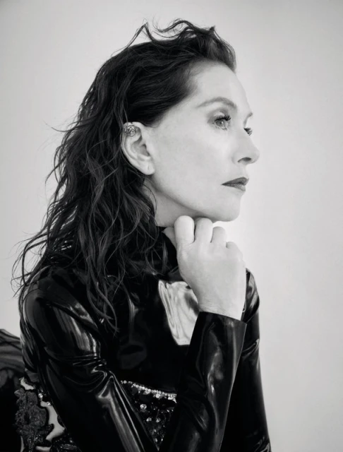 Black and white portrait of an adult woman with dark hair, styled with an ear cuff, wearing a black, high-necked, reflective top, and posing indoors.