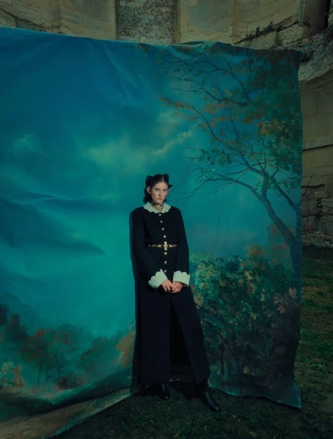 Woman in long dark coat posing in front of a backdrop with a painted nature scene, standing on grass, fashion photography.