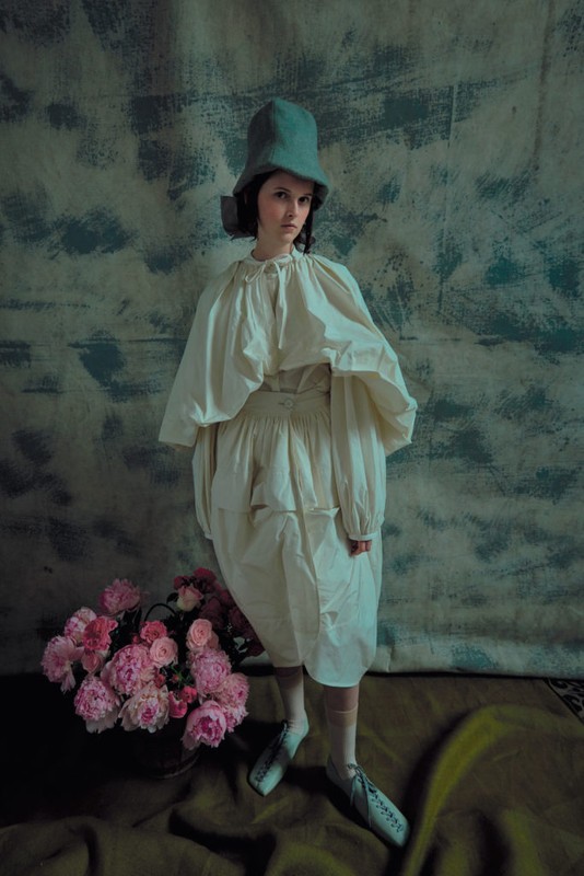 A woman in a vintage dress and hat stands near a bucket of pink flowers in a studio, creating a whimsical and nostalgic fashion portrait.