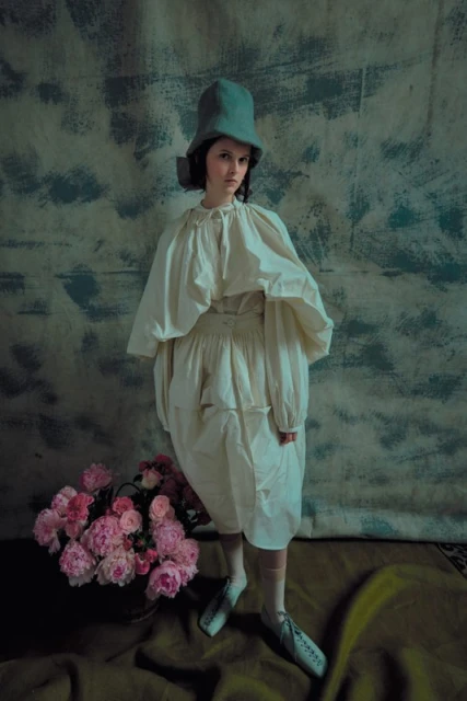 A woman in a vintage dress and hat stands near a bucket of pink flowers in a studio, creating a whimsical and nostalgic fashion portrait.