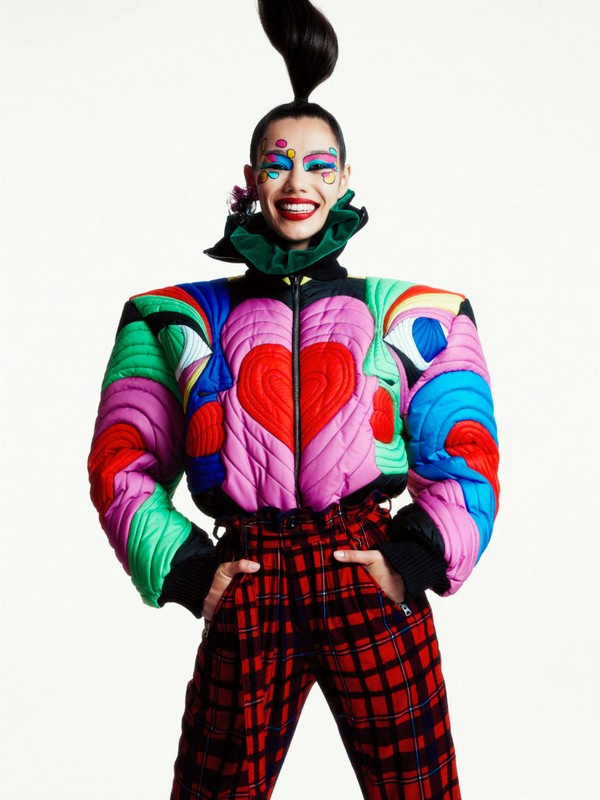 A smiling adult female in a colorful fashion jacket and plaid pants stands in a studio setting. The model is shot at eye-level.