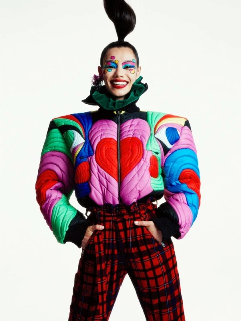 A smiling adult female in a colorful fashion jacket and plaid pants stands in a studio setting. The model is shot at eye-level.