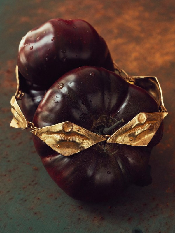 Close-up still life of two ripe, dark purple tomatoes adorned with an elaborate gold necklace, placed on a mottled teal and brown surface.