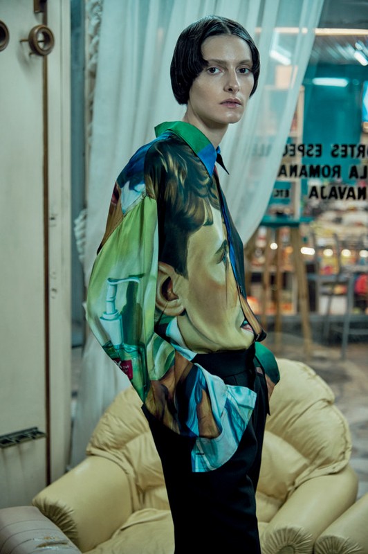 Fashion shot of a woman wearing a silk shirt with printed faces standing in front of a chair and window with visible shop interior.