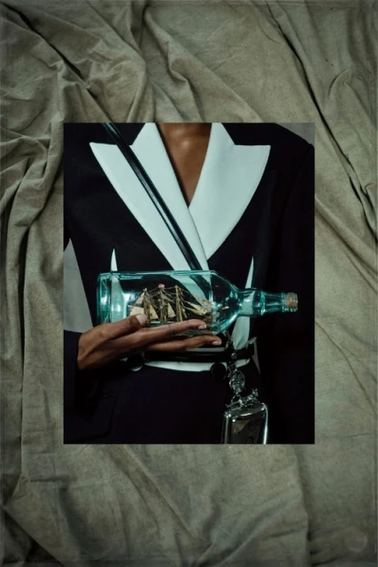 Person in blazer holding a ship-in-a-bottle against a draped fabric backdrop, creating an unusual and eye-catching presentation.