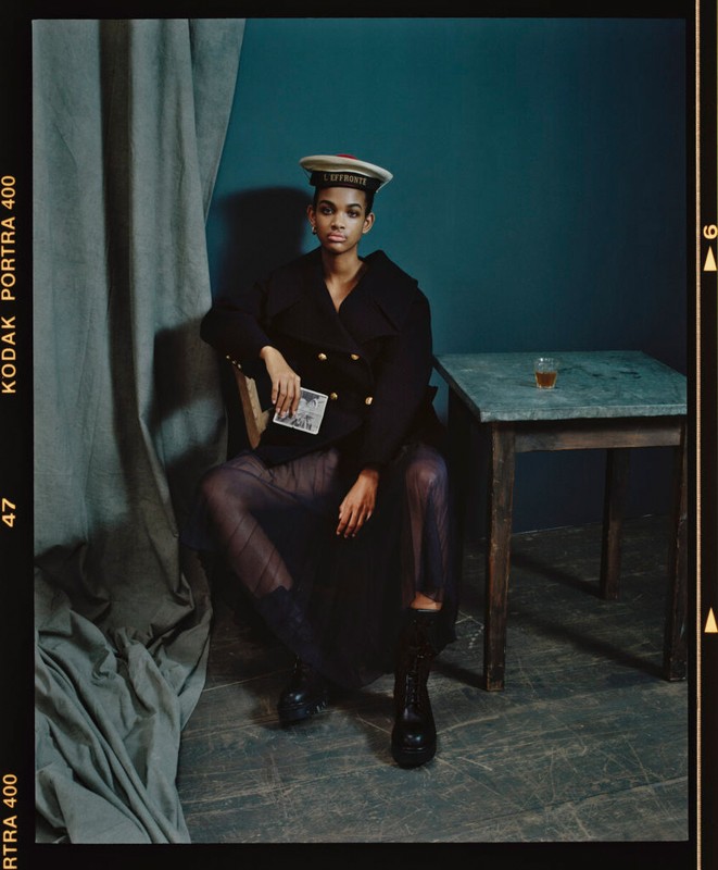 Portrait of a woman in dark clothing and a nautical hat seated indoors, holding a card, next to a table with a glass.