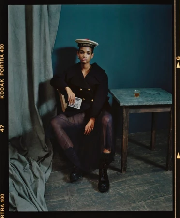 Portrait of a woman in dark clothing and a nautical hat seated indoors, holding a card, next to a table with a glass.