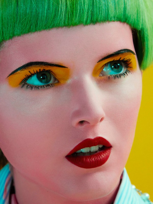 Close-up portrait of a woman with bold makeup, including yellow eyeshadow, red lipstick, green blunt-cut hair, and light skin, on a yellow background.