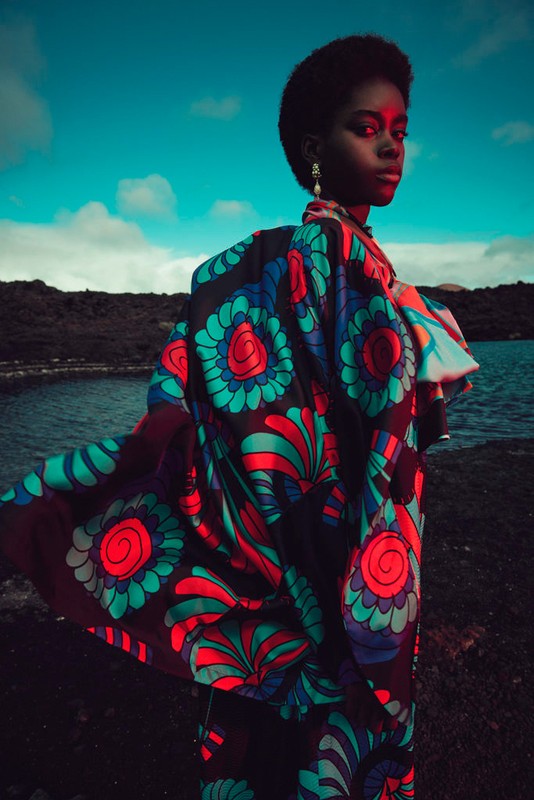 Outdoor fashion shot of an African woman in a vibrant floral-print cape by a lake with an expressive gaze, emphasizing texture and color.