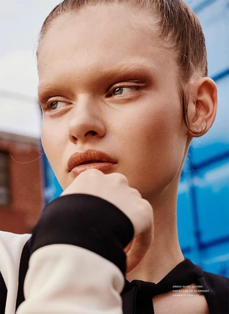 Close-up portrait of a fair-skinned adult female model with neutral expression, slicked-back hair, and a black and white top in an urban setting.