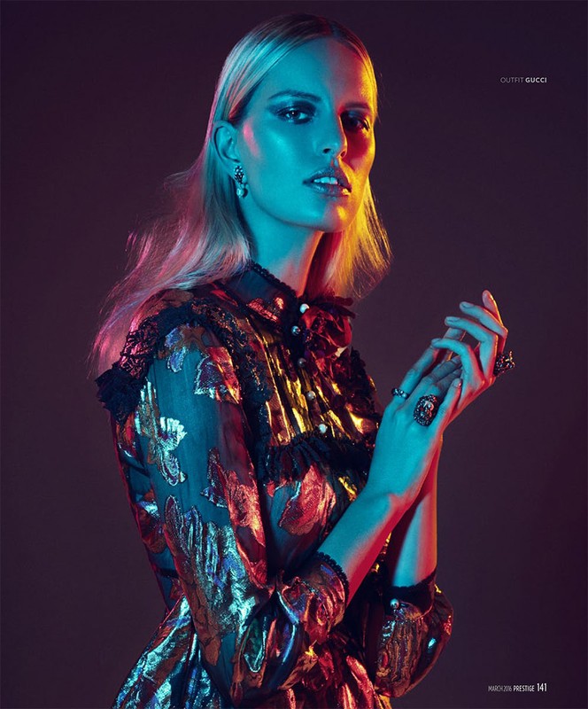 Fashionable woman in shimmering Gucci dress, standing indoors with dramatic lighting. Statement rings and earrings accessorize the look.