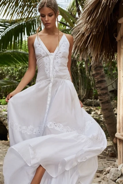 Woman in long white dress posing outdoors in a tropical setting with palm trees and thatched roof structure, fashion and travel photography.