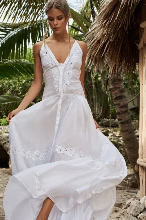 Woman in long white dress posing outdoors in a tropical setting with palm trees and thatched roof structure, fashion and travel photography.