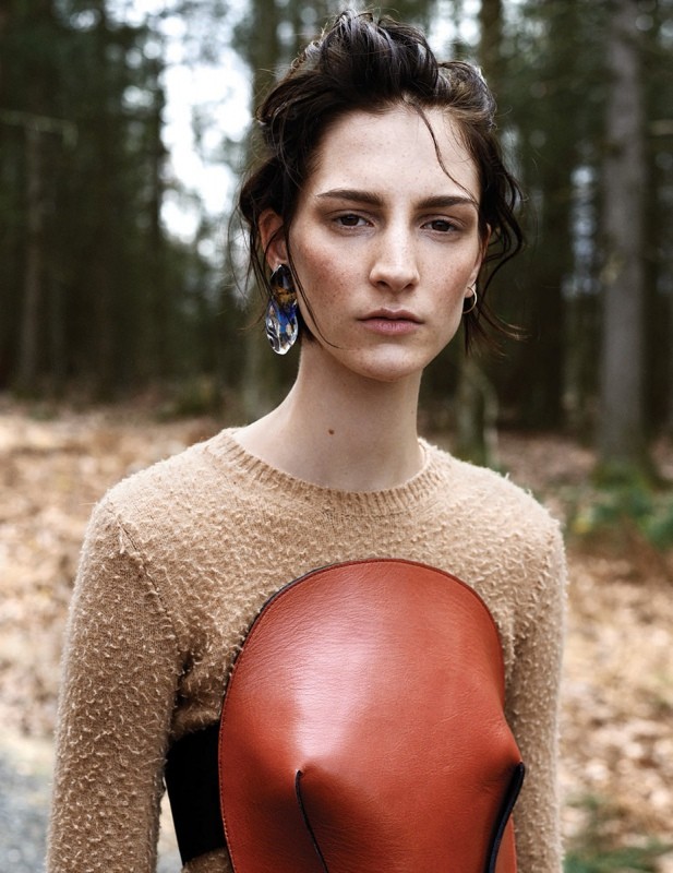 Portrait of a young woman with dark hair wearing a beige sweater and a red leather piece against a blurred forest background.