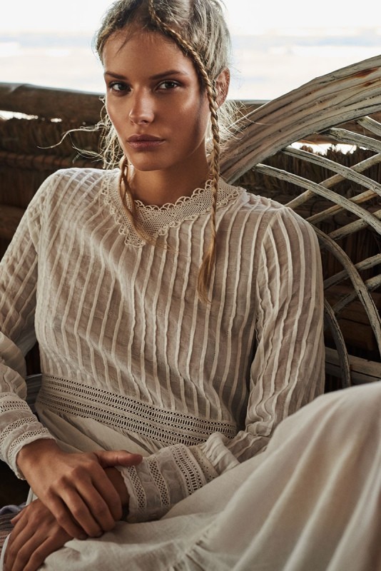 Close-up portrait of a female model wearing a white lace-trimmed blouse with her hair in braids against a textured woven background