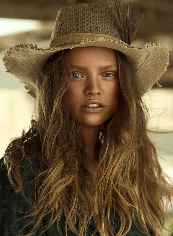 Close-up portrait of a tan-skinned woman with long wavy hair wearing a straw hat with a feather accent and green shirt. Studio lighting.