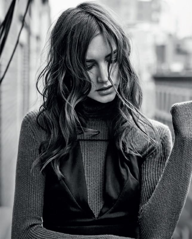 Monochrome portrait of a woman with wavy long hair, wearing a turtleneck and dress, in front of an urban background.