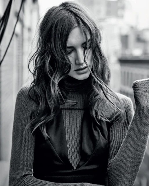 Monochrome portrait of a woman with wavy long hair, wearing a turtleneck and dress, in front of an urban background.