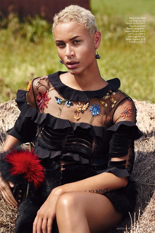 Portrait of a young woman with short blonde hair, adorned with floral dress, seated outdoors with blurred background, fashion editorial.