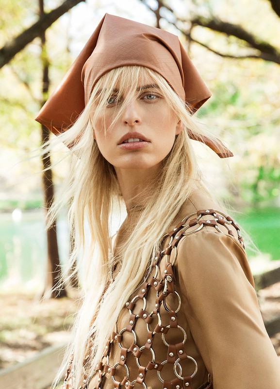 Woman with light hair and a brown headscarf poses outdoors, wearing a top made of linked metal rings and a beige shirt.