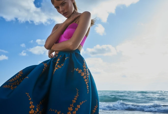 Woman in a pink top and blue skirt stands on the beach with the ocean in the background on a sunny day with blue skies and clouds.