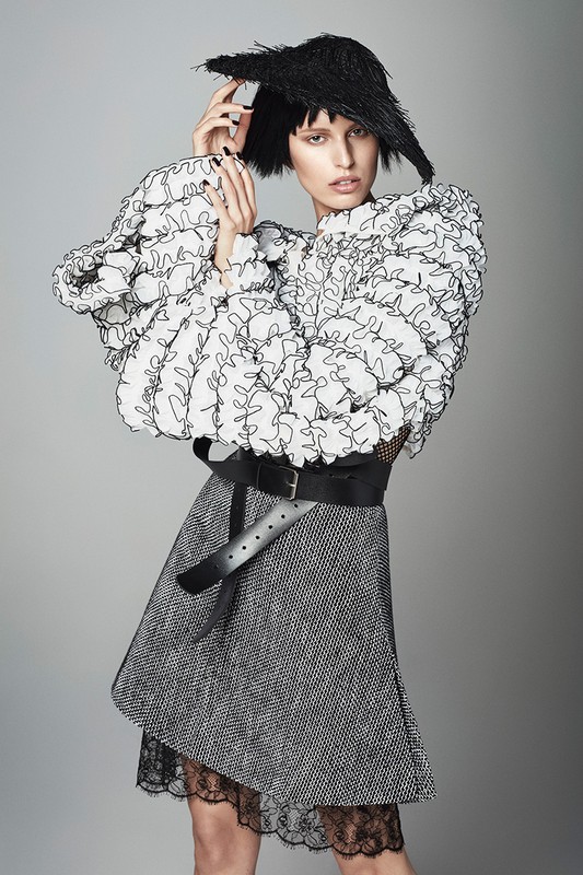 Fashion model poses in a white ruffled top, gray skirt with lace trim, wide black belt, and unique black headwear against a gray backdrop.