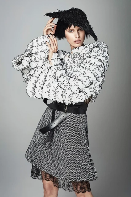 Fashion model poses in a white ruffled top, gray skirt with lace trim, wide black belt, and unique black headwear against a gray backdrop.
