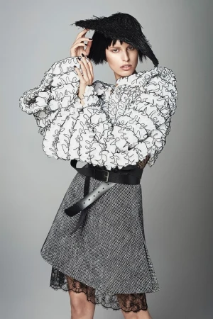 Fashion model poses in a white ruffled top, gray skirt with lace trim, wide black belt, and unique black headwear against a gray backdrop.