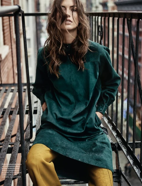 Fashion model in emerald green tunic and gold pants poses on a fire escape with brick buildings in the background, medium shot.