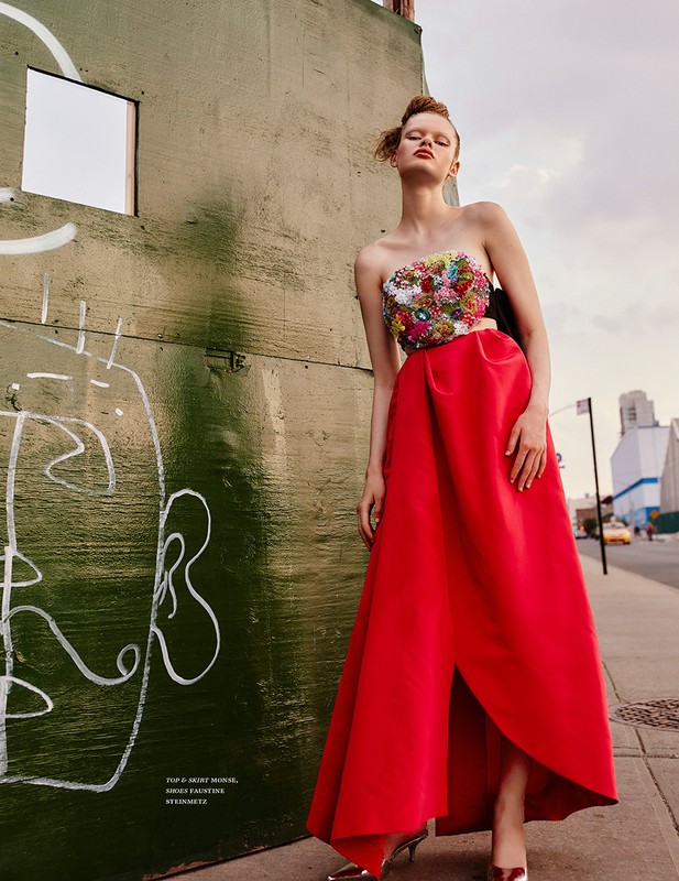 Model standing on a city sidewalk wearing a strapless floral embellished top and a long red skirt next to a green wall.