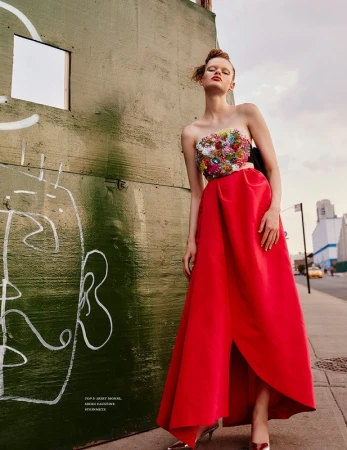 Model standing on a city sidewalk wearing a strapless floral embellished top and a long red skirt next to a green wall.