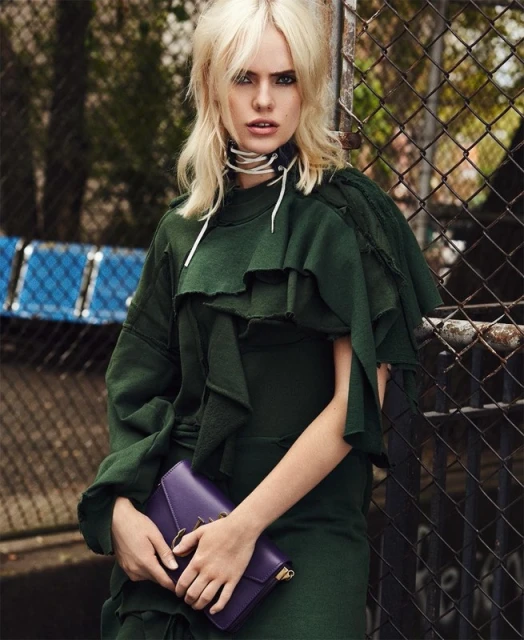 Fashionable woman with blonde hair poses outdoors wearing a green dress and purple purse, standing near a chain-link fence.