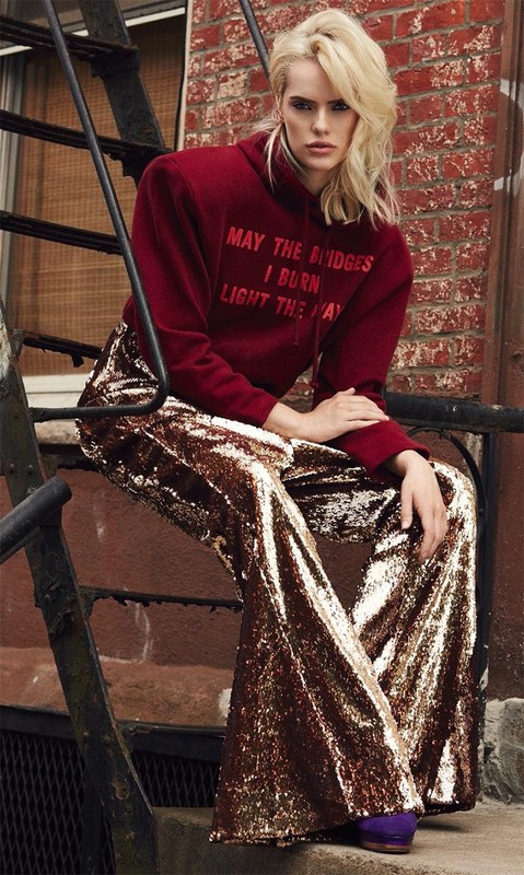 Blonde model in maroon hoodie and sequin bell-bottoms poses on metal steps outside, showcasing fashion and urban style with neutral backdrop.