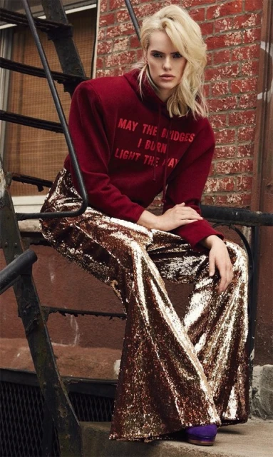 Blonde model in maroon hoodie and sequin bell-bottoms poses on metal steps outside, showcasing fashion and urban style with neutral backdrop.