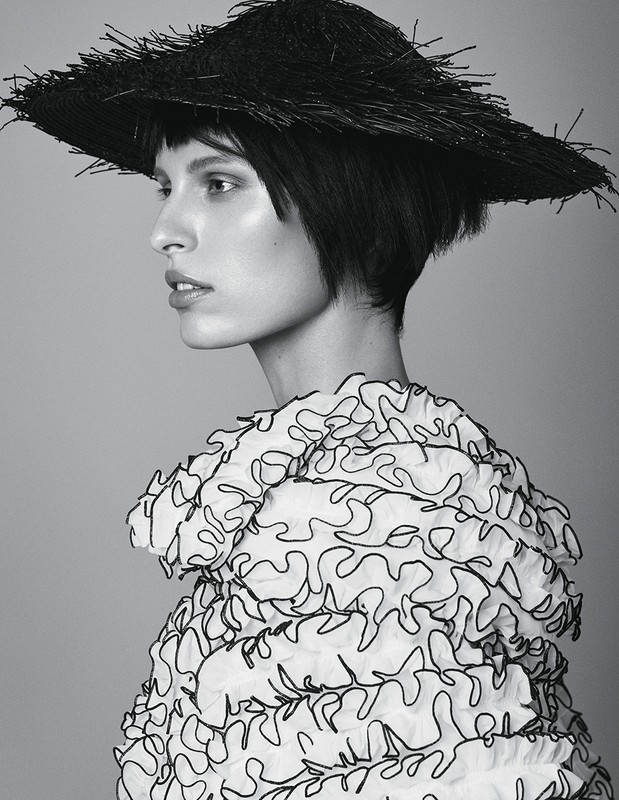 Monochrome fashion portrait of a fair-skinned woman with a black bob, wearing a straw hat and ruffled white top against a grey backdrop.