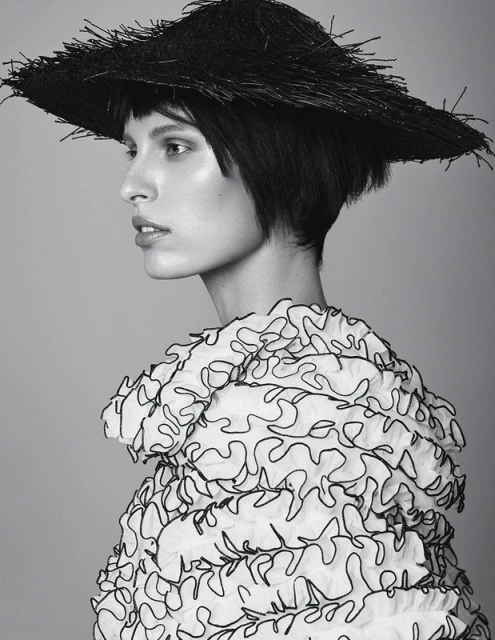 Monochrome fashion portrait of a fair-skinned woman with a black bob, wearing a straw hat and ruffled white top against a grey backdrop.