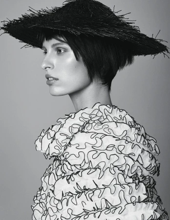 Monochrome fashion portrait of a fair-skinned woman with a black bob, wearing a straw hat and ruffled white top against a grey backdrop.