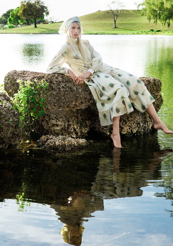 Model wearing a patterned cream dress and headscarf seated on a mossy rock by a lake with bare feet touching the water