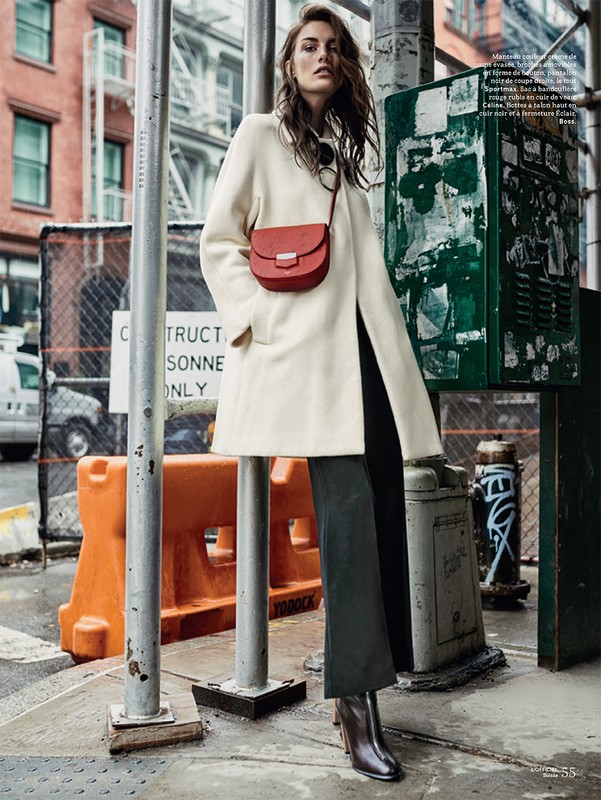 Fashionable woman in a cream-colored coat and dark trousers stands on a city street with an orange crossbody bag and ankle boots.