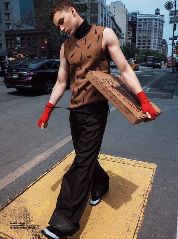 A young adult male walks on a city street wearing a sweater vest and carrying a pizza box. Urban fashion photography. 