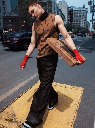 A young adult male walks on a city street wearing a sweater vest and carrying a pizza box. Urban fashion photography. 